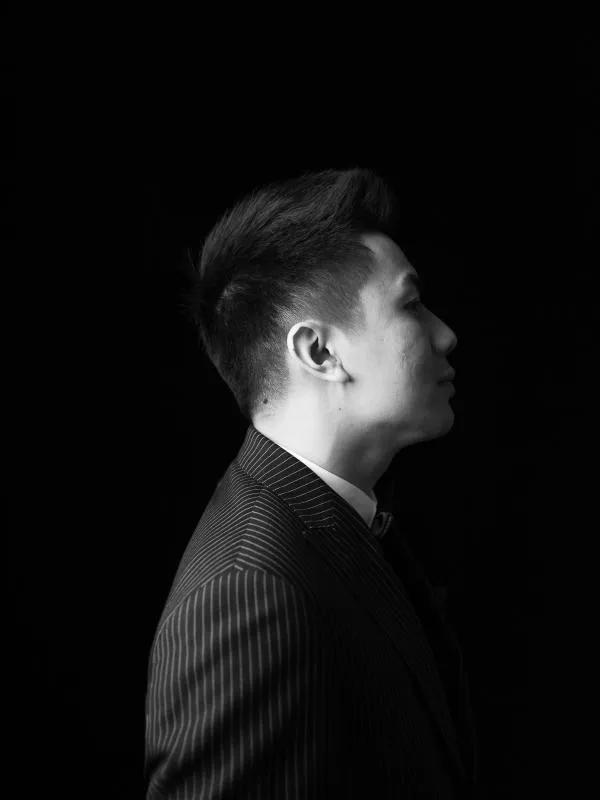 Black-and-white photo of an Asian man turned to the side wearing a striped black suit.