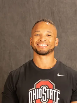 OPEEP Advisory Council member and Lecturer in Department of Sociology at OSU smiles while standing for a professional headshot against a dark gray background while wearing a black t-shirt with a white-and-red OSU logo and a white Nike swoosh on the front.